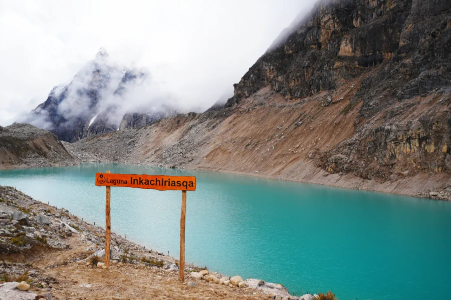 Close-up of a wooden trail sign beside Incachiriasqa Lake with mountains in the background | Conde Travel Adventures